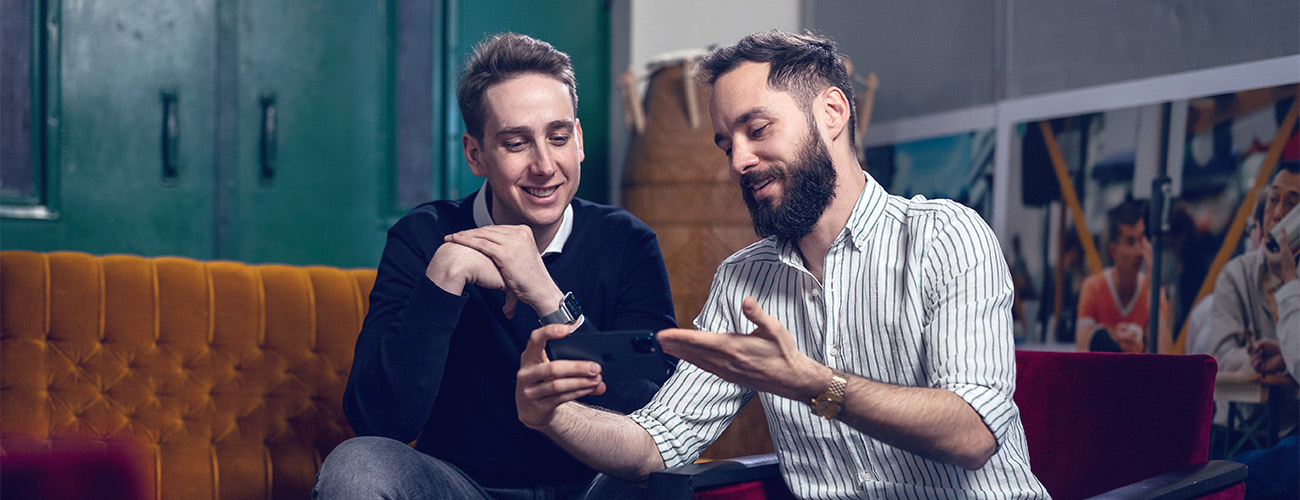 Dva moška sedita na kavču in se z nasmehom pogovarjata, medtem ko en izmed njiju drugemu nekaj kaže na pametnem telefonu.