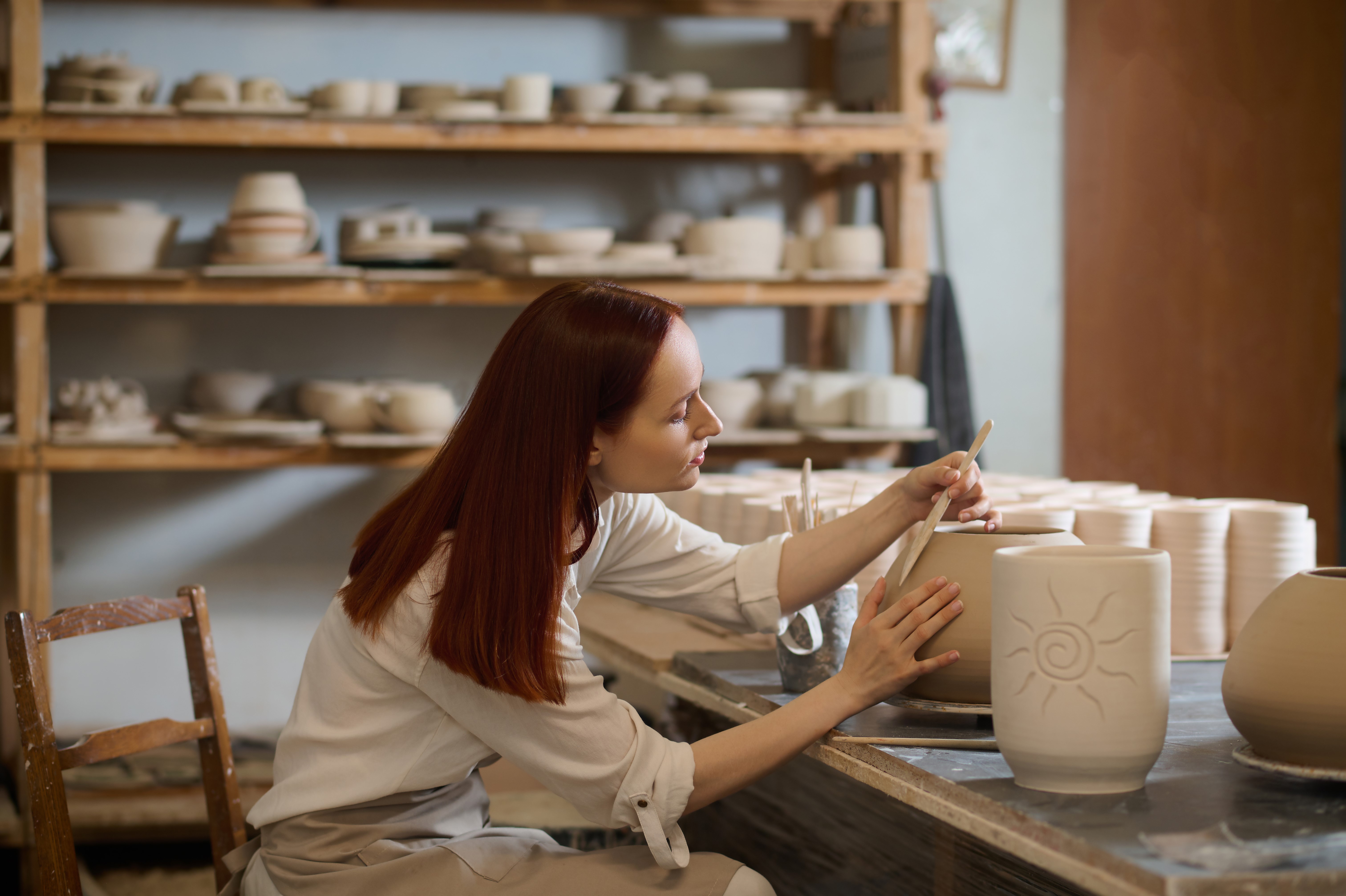 Na sliki je ženska ki natančno oblikuje glineno posodo v lončarski delavnici obdani s policami keramike
