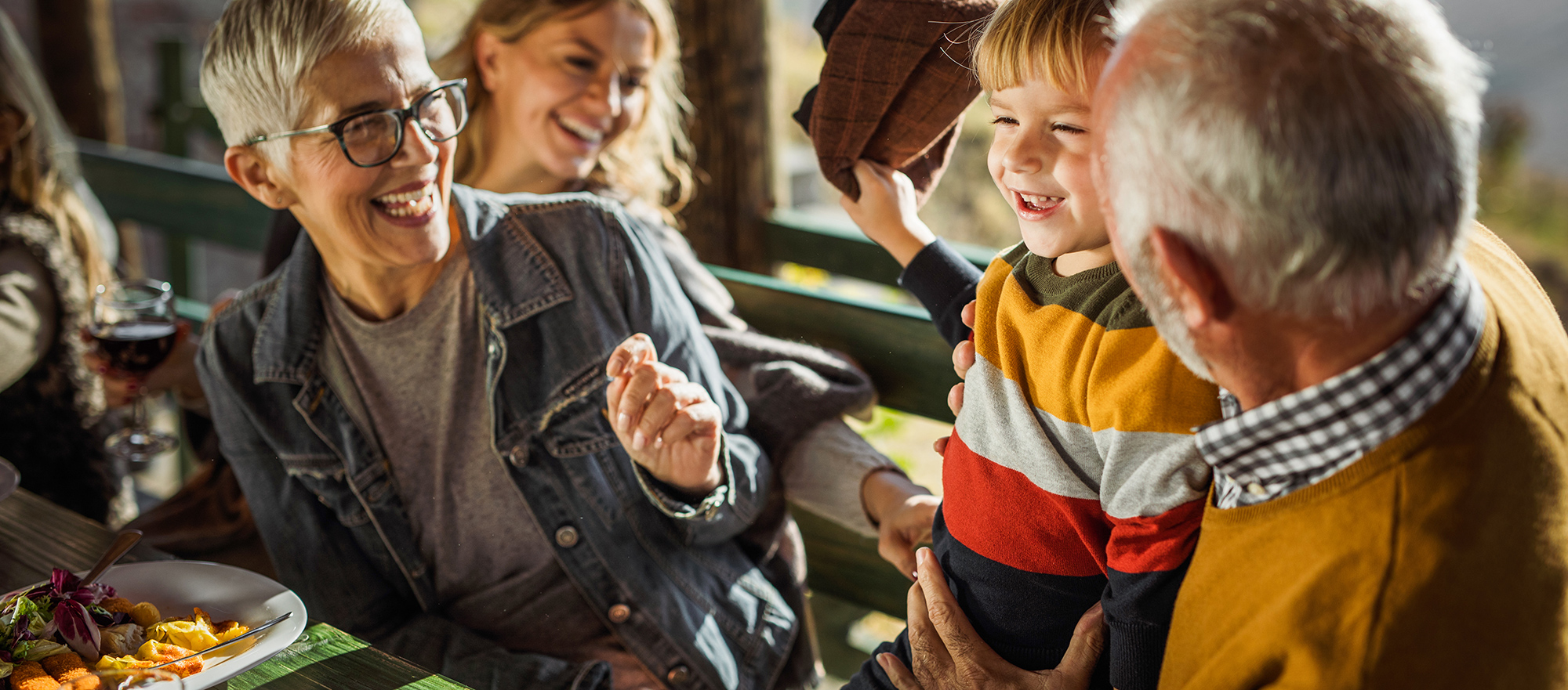 Večgeneracijska družina se smeji in uživa skupne trenutke za mizo na prostem ob sončnem dnevu