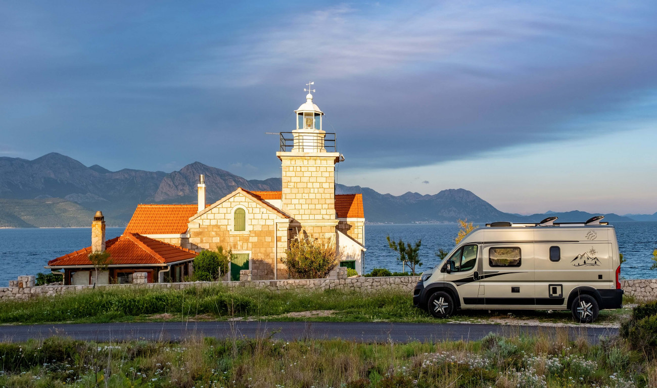 Na sliki je svetilnik z rdečo streho ob morju z avtodomom parkiranim ob cesti in gorami v ozadju