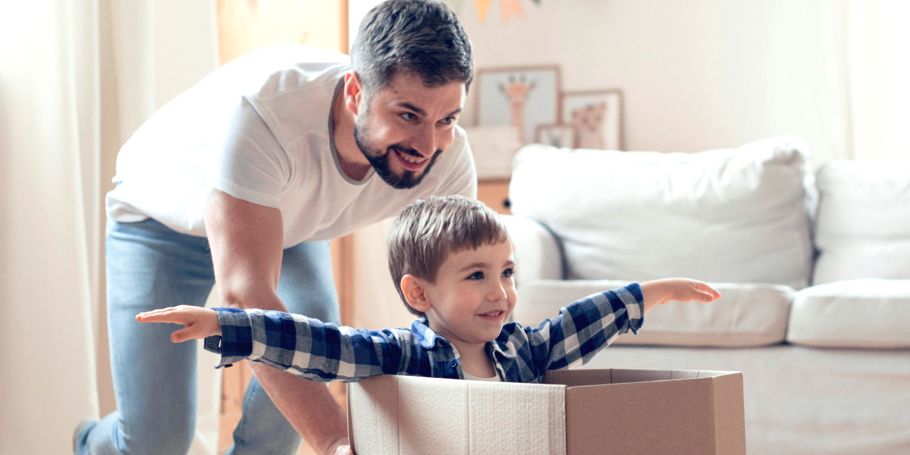 Otrok sedi v kartonasti škatli na tleh, oče od zadaj potiska škatlo naprej