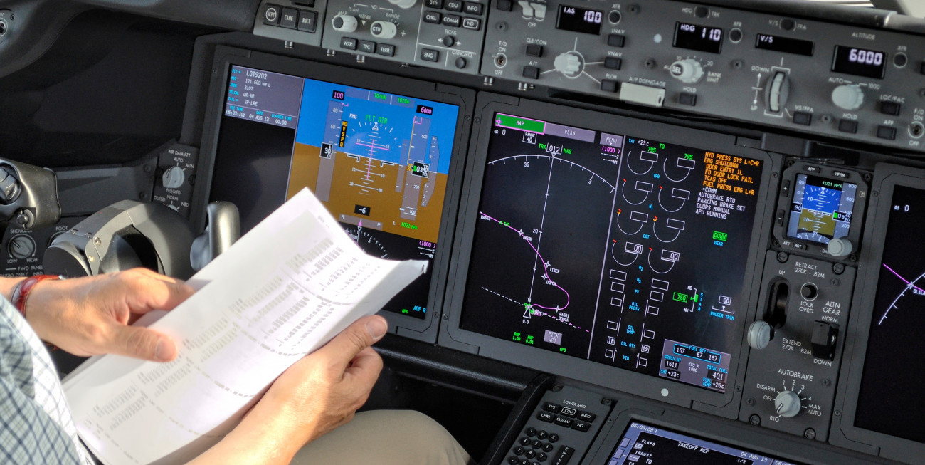 Pilot v pilotski kabini drži navigacijski seznam pred sodobnimi zasloni z letalskimi instrumenti in letalsko potjo