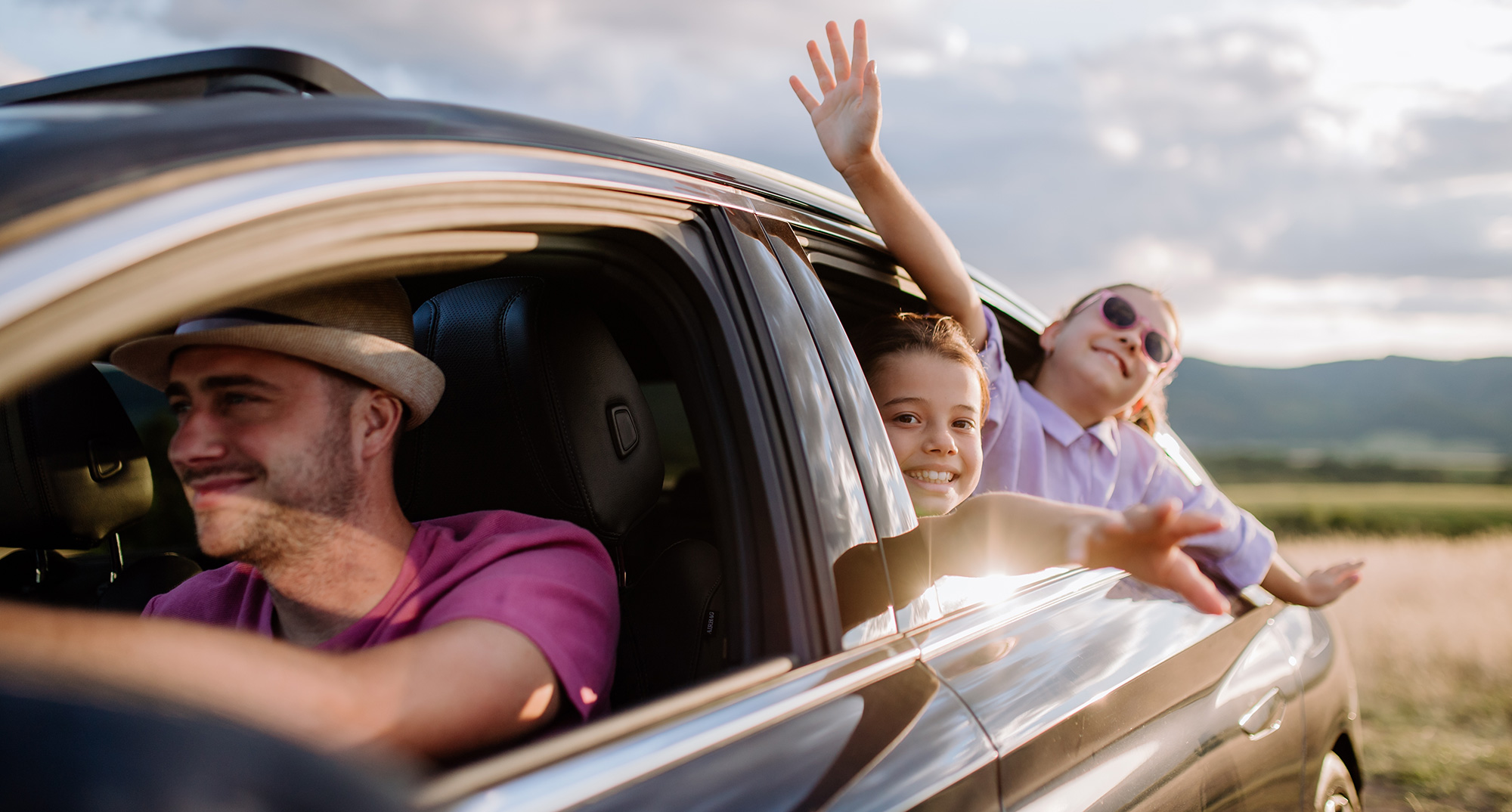 Nasmejan moški vozi avto medtem ko se otroka skozi okno veselita in mahata med vožnjo po podeželju