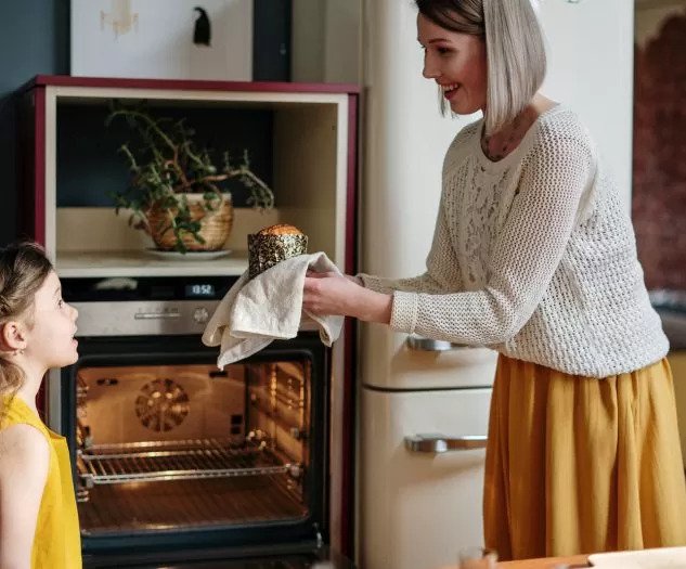 Ženska z nasmehom drži sveže pečen kruh pred odprto pečico in ga ponosno ponudi deklici ki jo opazuje v domači kuhinji