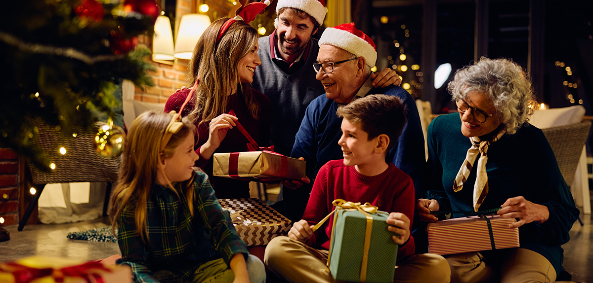 Družina v prazničnem vzdušju sedi ob božičnem drevesu in si z nasmehi izmenjuje darila
